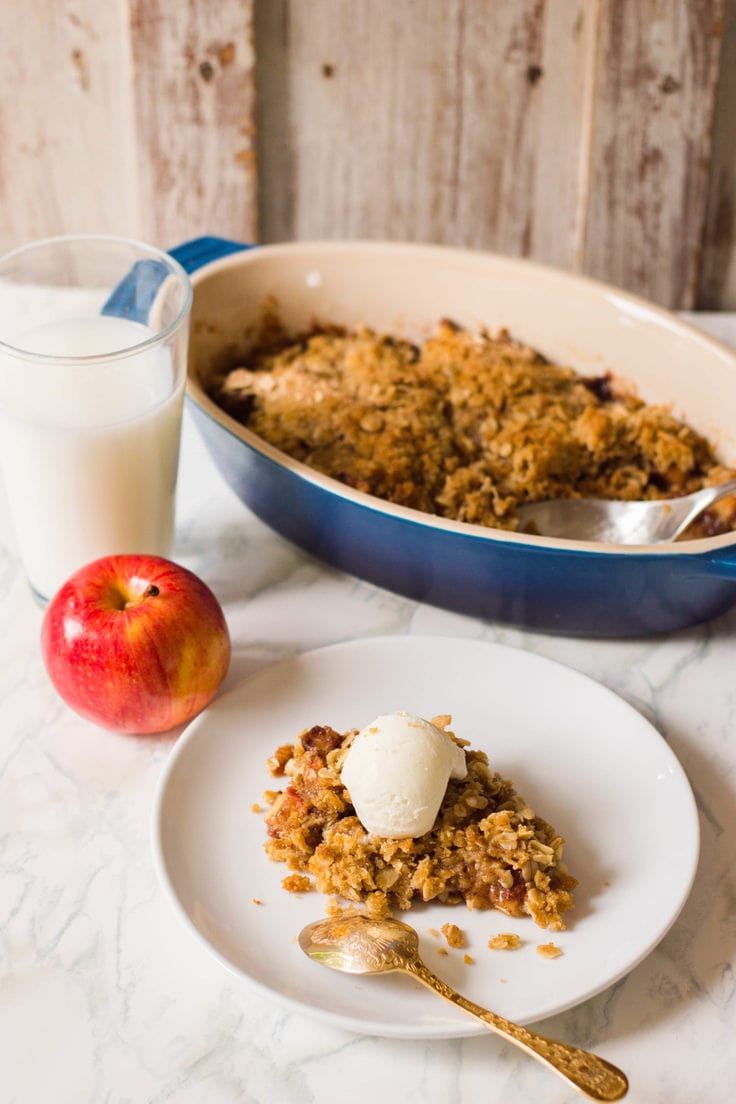 A slice of apple crisp topped with a scoop of vanilla ice cream on a white plate beside the casserole dish.