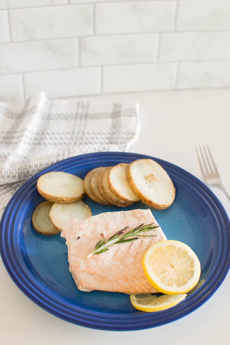 Baked salmon filet topped with rosemary and lemons, with a side of potatoes on a bright blue plate.