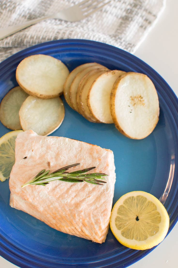 Baked salmon filet topped with rosemary and lemons, with a side of potatoes on a bright blue plate.