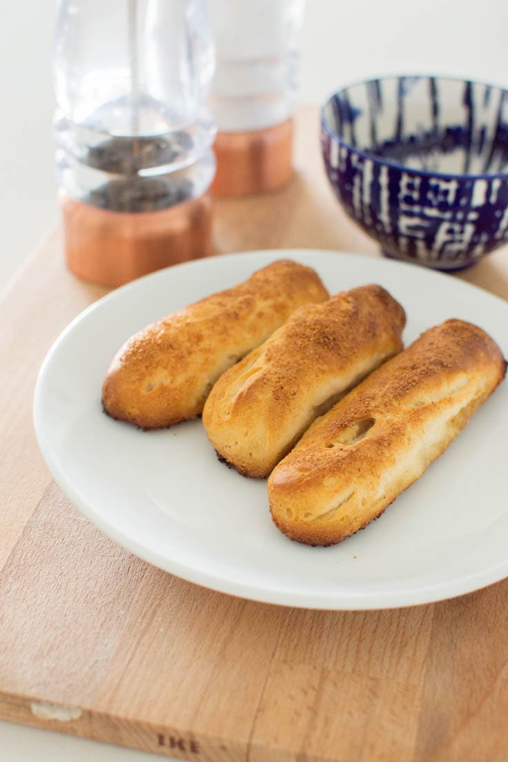 Three freshly baked garlic breadsticks on a white plate, surrounded by a tie-dye blue bowl and large salt and pepper shakers