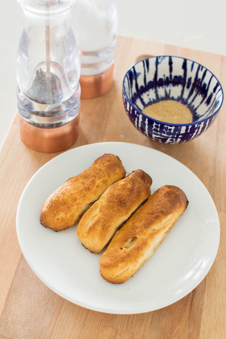 Three freshly baked garlic breadsticks on a white plate, surrounded by a tie-dye blue bowl and large salt and pepper shakers