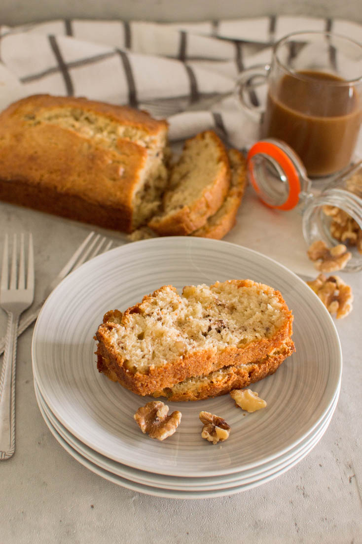 Two slices of walnut loaf piled on top of each other on a grey plate, surrounded by forks, walnuts, the entire loaf and a mason jar full of coffee.