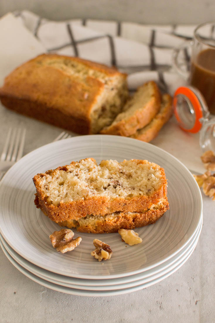 Two slices of walnut loaf piled on top of each other on a grey plate, surrounded by forks, walnuts, the entire loaf and a mason jar full of coffee.