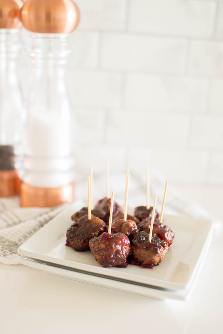 Meatballs with a toothpick in each one, sitting on a white square plate with a salt and pepper shaker in the background