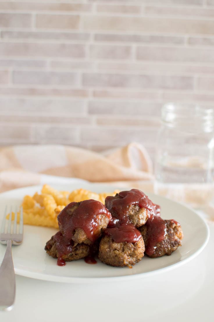 Beef meatballs topped with a cranberry sauce on a white plate with a side of fries, with a peach plaid napkin in the background