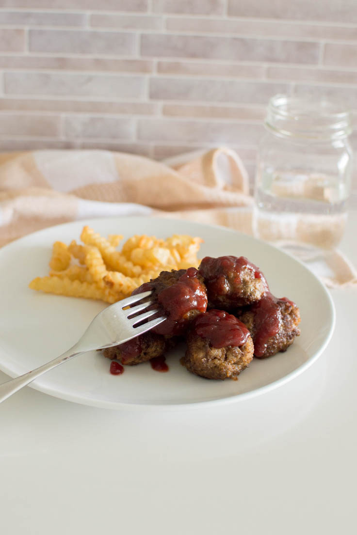 Beef meatballs topped with a cranberry sauce on a white plate with a side of fries, with a peach plaid napkin in the background, with a fork inserted into one of the meatballs