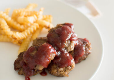 A closeup of homemade beef meatballs, with a cranberry sauce topping on a white plate with a side of fries