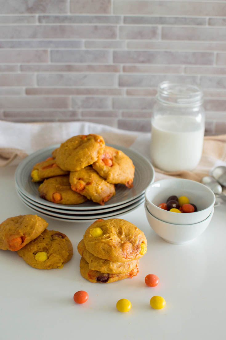 Homemade pumpkin cookies sitting on a stack of grey plates with a mason jar of milk, surrounded by additional candies and cookies