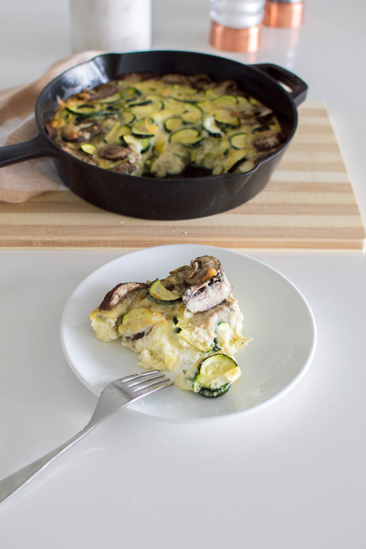 A slice of a vegetable frittata on a white plate with a skillet and cutting board in the background