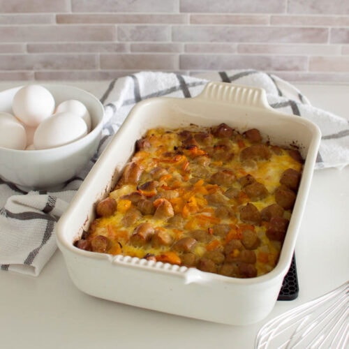 Hash brown breakfast casserole, topped off with orange bell pepper and sausages in a white casserole pan, with a grey and white napkin and a bowl of eggs in the background