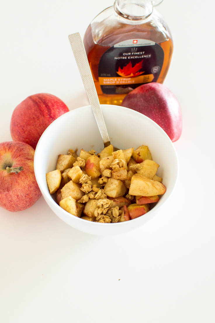 Cooked apples in a small white bowl with a spoon, surrounded by apples and maple syrup