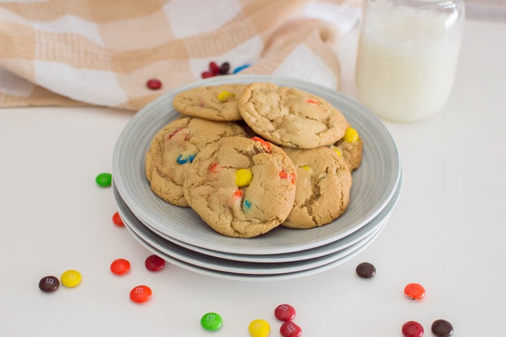 A plate of M&M cookies piled on a stack of grey plates, surrounded by milk chocolate M&Ms