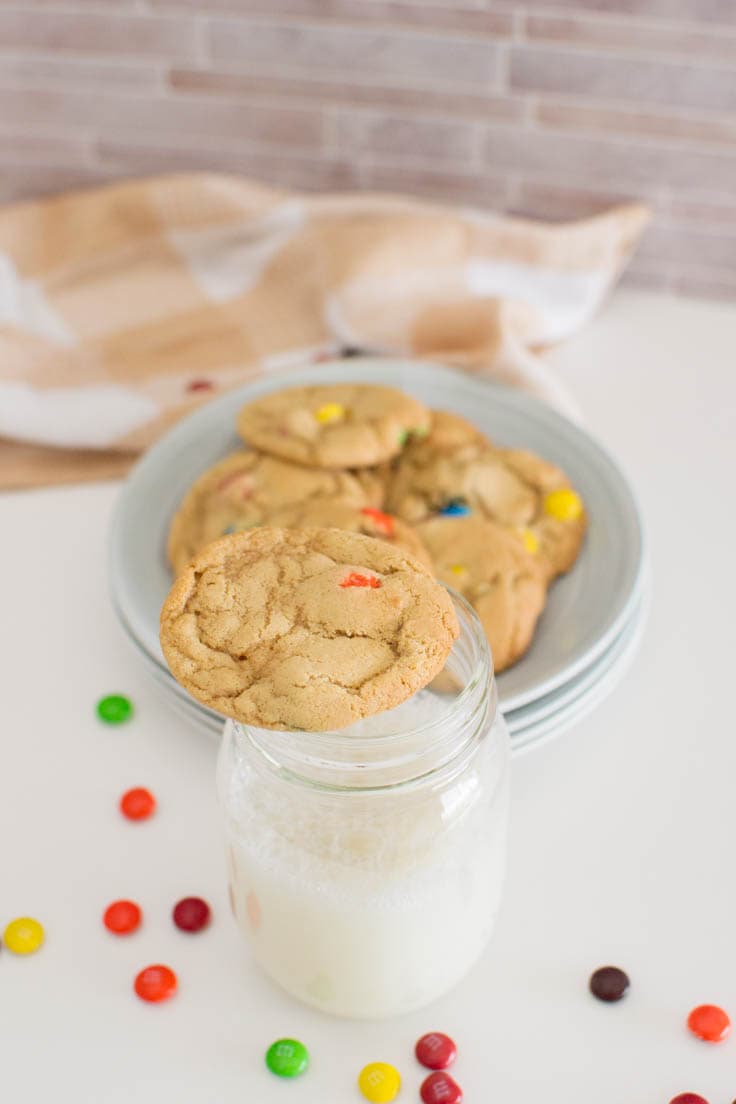 An M&M cookie sits on top of a tall glass of milk with a plate of cookies in the background.