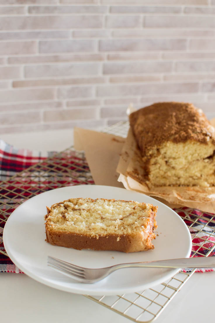 A slice of Cinnamon Bread sits on a white plate, with the loaf in the background, on top of a plaid place mat