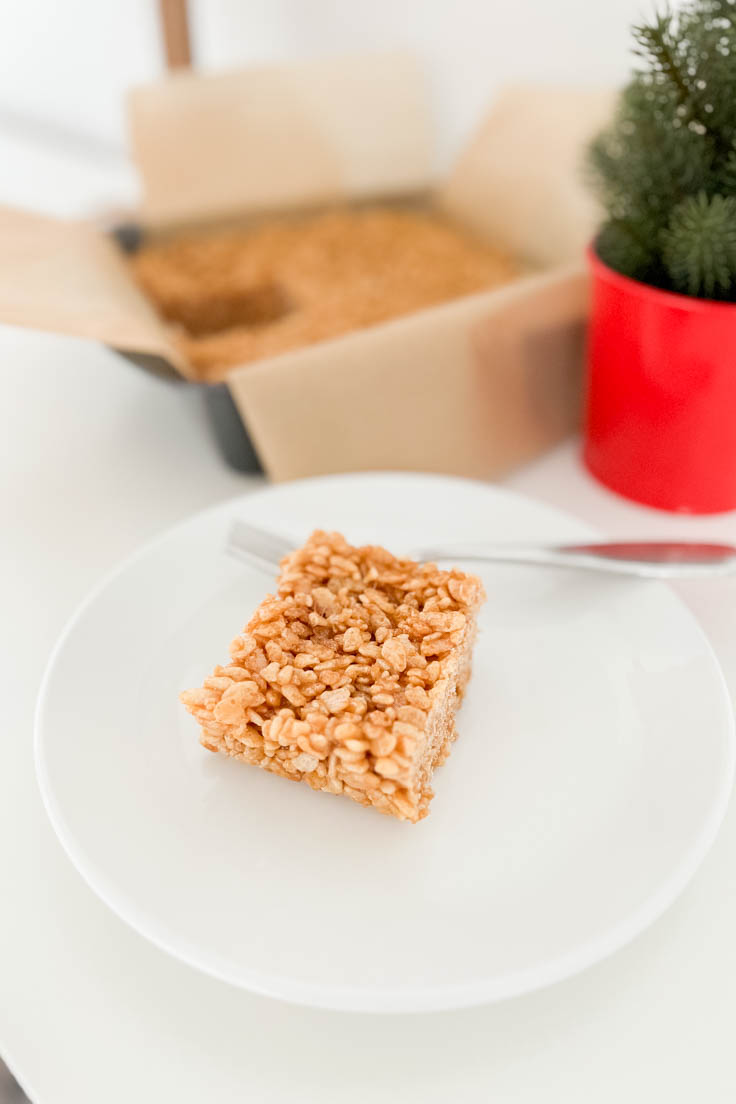 A gingerbread-flavored rice cereal treat on a white plate, with a full pan of Gingerbread rice cereal treats in the background