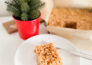 A gingerbread-flavored rice cereal treat on a white plate, with a full pan of Gingerbread rice cereal treats in the background