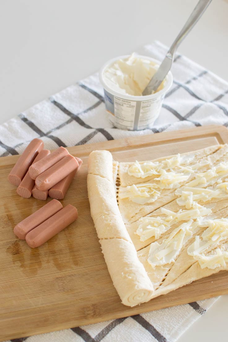 Adding cream cheese and shredded cheese on canned croissant dough, a pile of hot dog halves sit next to the dough