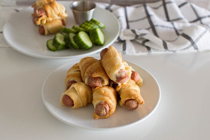 A pile of Air Fryer Pigs in a Blanket on a white plate, ready to be eaten, with some more in the background along with dip and slices of cucumber