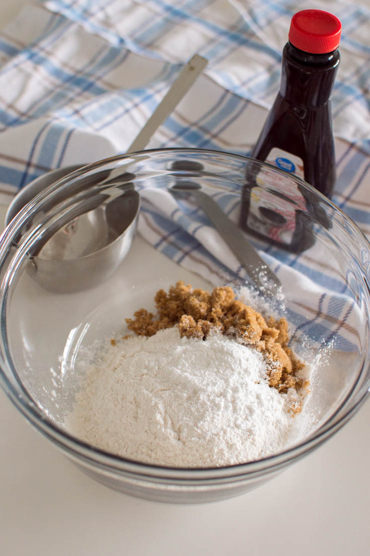 Adding dry ingredients to make Brown Sugar Pancakes to a large glass bowl, with a plaid tea towel in the background
