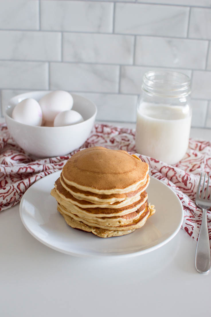 A stack of Brown Sugar Pancakes on a red tea towel with a glass of milk and a bowl of eggs in the background