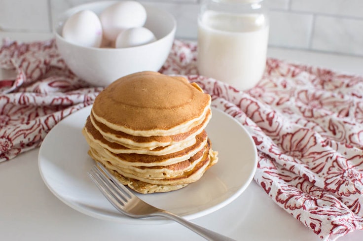 A stack of pancakes on a white plate, with a fork beside it. A red tea towel sits in the background, along with a mason jar glass of milk and a bowl of eggs