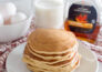 A close-up shot of a stack of pancakes, sitting on a white plate, with maple syrup, a tall glass of milk, and a bowl of eggs in the background
