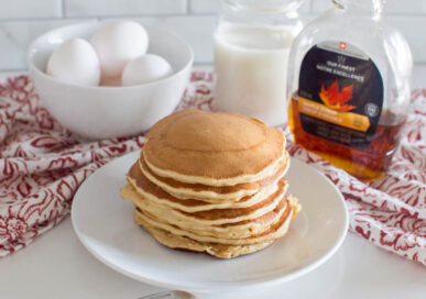 Stacked brown sugar pancakes on a white plate, with eggs, milk and maple syrup in the background