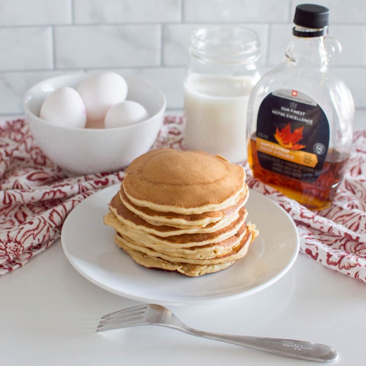 Stacked brown sugar pancakes on a white plate, with eggs, milk and maple syrup in the background