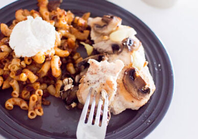 A piece of oven-baked chicken on a fork, laying on a grey plate, served with macaroni pasta