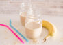 Two mason jars filled with peanut butter oatmeal smoothie, paired with two silicone straws and a banana sits in the background