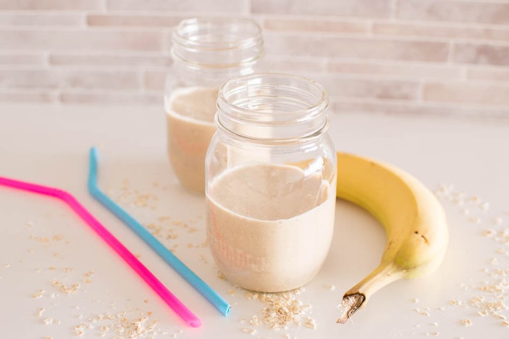 Two mason jars filled with peanut butter oatmeal smoothie, paired with two silicone straws and a banana sits in the background