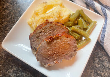 sliced of meatloaf on a white plate with green beans mashed potatoes and a blue and white napkin