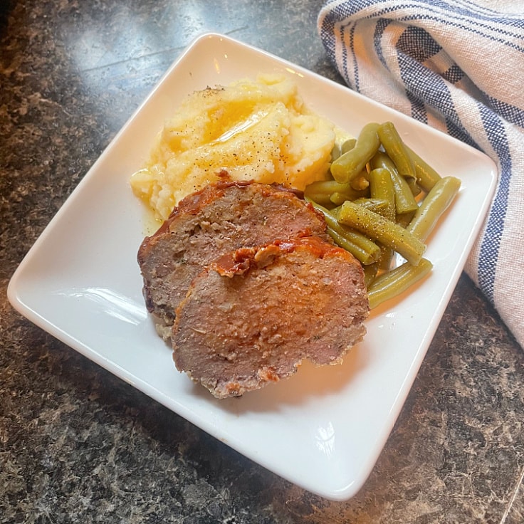 sliced of meatloaf on a white plate with green beans mashed potatoes and a blue and white napkin