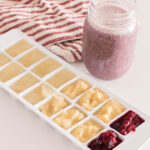 Almond milk, mashed bananas, and mashed berries frozen in a white ice cube tray, accompanied by a smoothie