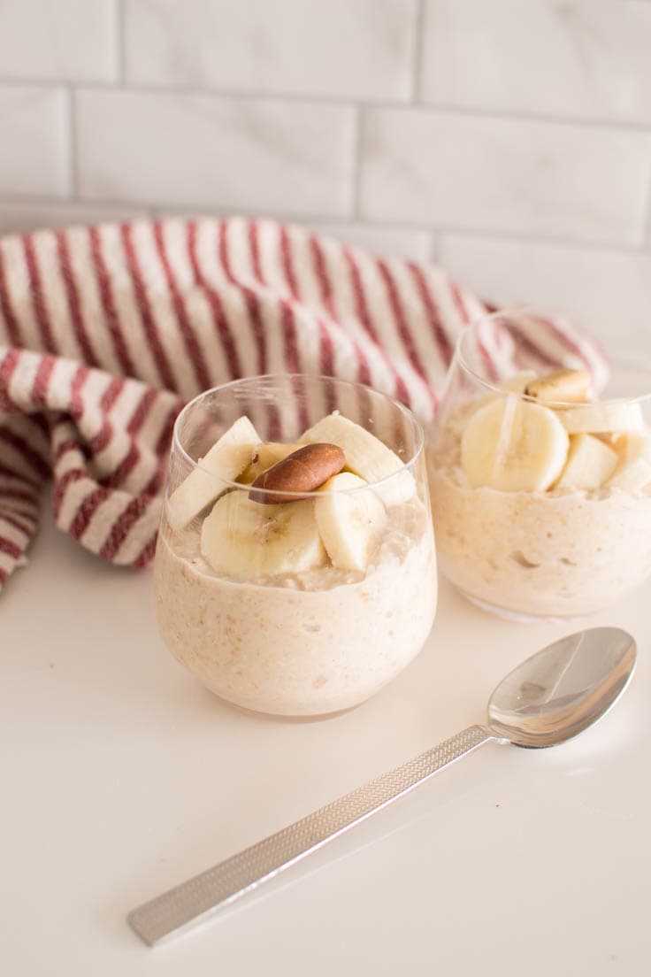 Peanut Butter Overnight Oats served in two small glasses, topped with banana slices and nuts. A red and white striped tea towel sits in the background