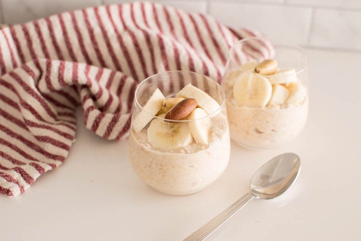 Peanut Butter Overnight Oats served in two small glasses, topped with banana slices and nuts. A red and white striped tea towel sits in the background