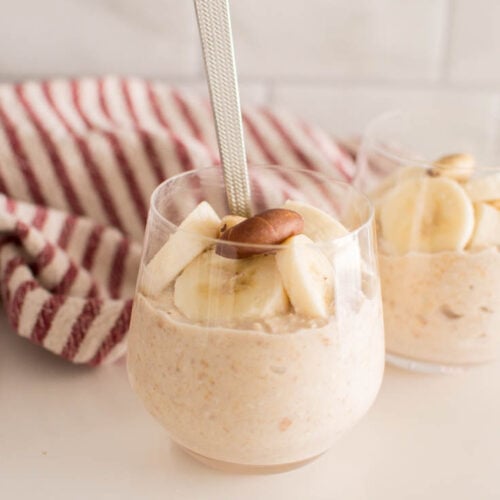 A close-up of a cup full of overnight oats, topped with bananas and nuts, with a spoon inserted into the cup