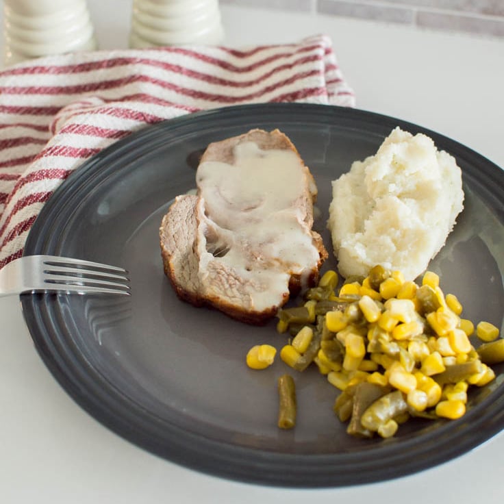 Slice of easy baked pork with gravy, side dishes, and a fork sit on a dark grey plate