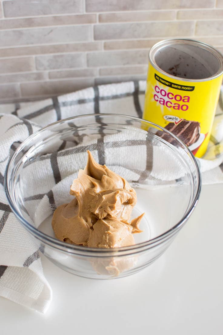 A glass bowl filled with creamy peanut butter and a can of cocoa powder in the background