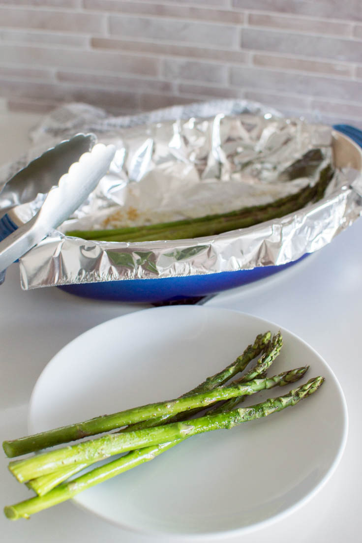 Freshly roasted asparagus on a small white plate and a tin foil-lined pan sits in the background