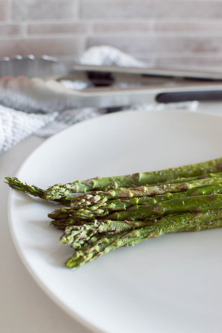 A close up shot of oven-roasted asparagus ready to be served on a white plate with tons in the background