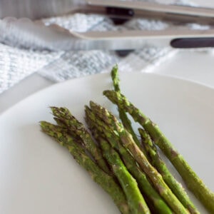 A close up shot of oven-roasted asparagus ready to be served on a white plate with tons in the background