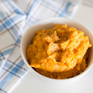 Aerial shot of our easy mashed sweet potatoes recipe, served in a white bowl with a plaid tea towel sitting beneath it