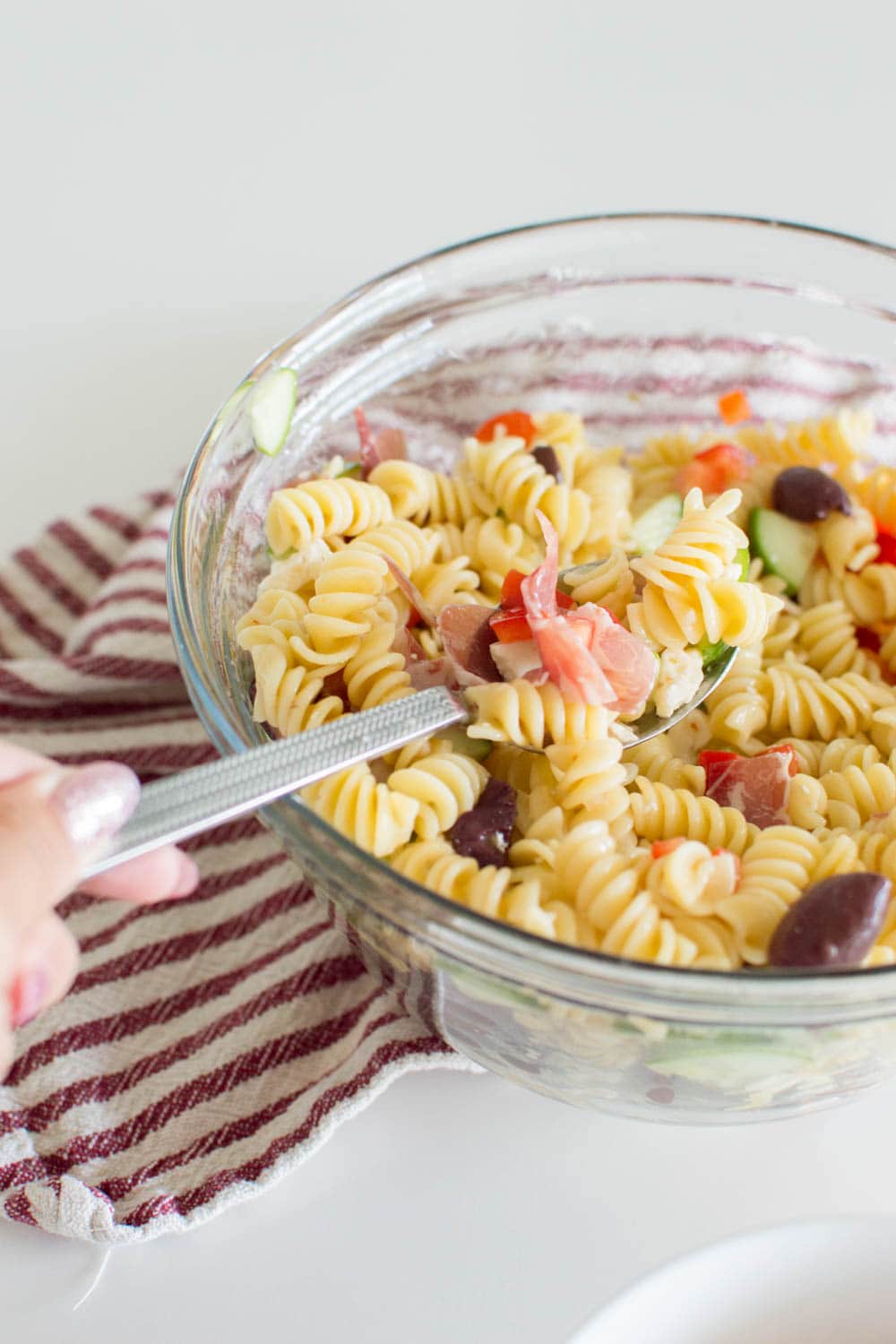 A spoon mixes pasta salad with Italian Dressing in a large glass bowl