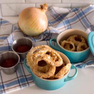 Homemade air fryer onion rings in turquoise bowls, surrounded by ketchup and a plaid tea towel