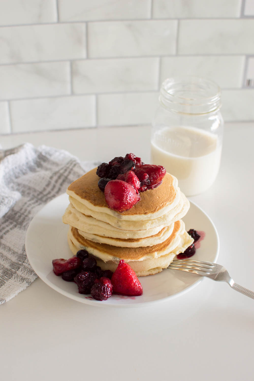 A stack of our delicious and easy homemade pancakes, topped with berry compote, sitting on a white plate and a fork