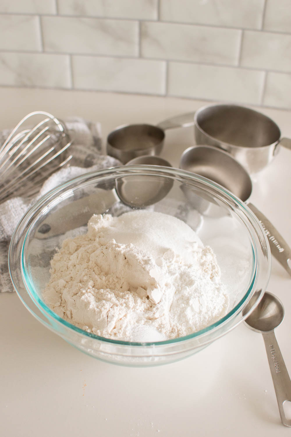 Dry ingredients for pancake batter in a glass bowl surrounded by measuring cups and a whisk