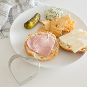 A cookie cutter slider with heart-shaped luncheon meat, open faced and accompanied by crackers, dip and a pickle
