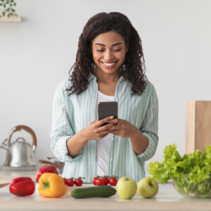 A woman looks as the Easy Recipe Depot meal planning app on her phone while standing at a kitchen counter covered in vegetables.