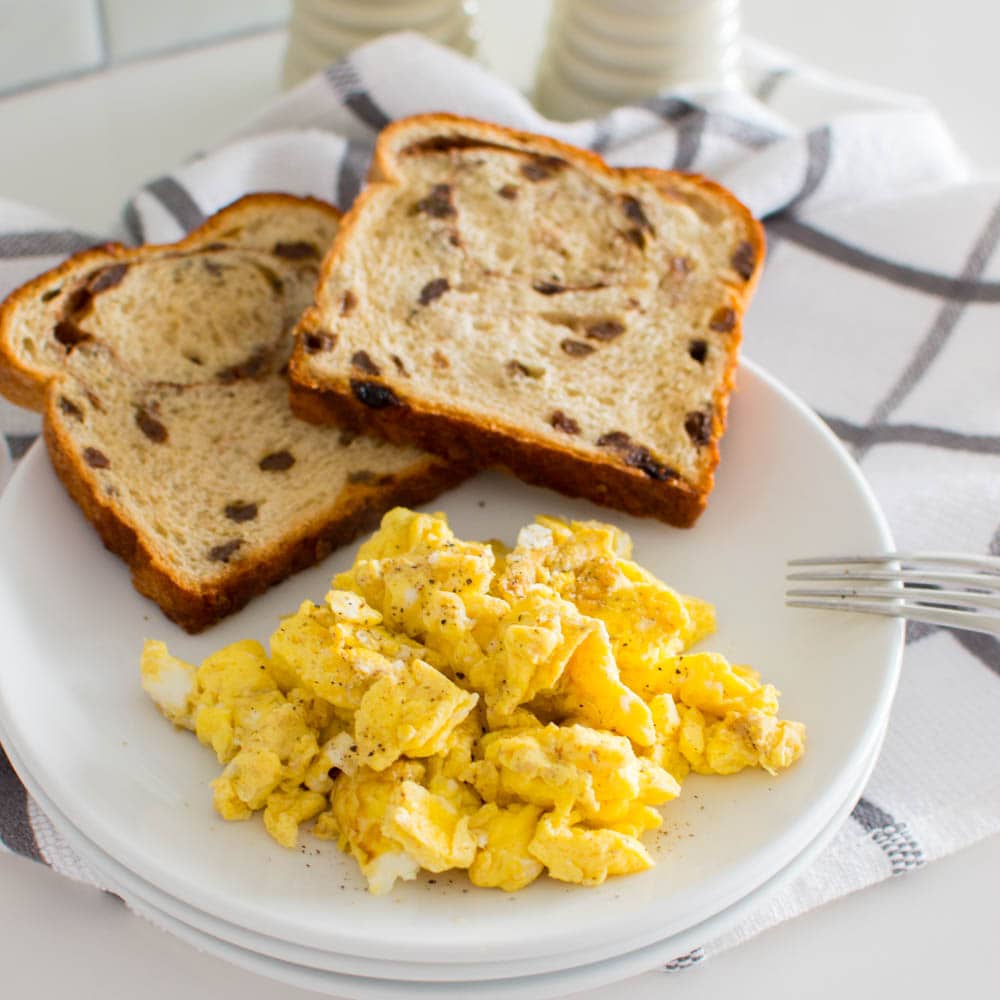 Easy Scrambled Eggs on a stack of white plates, accompanied by raisin bread